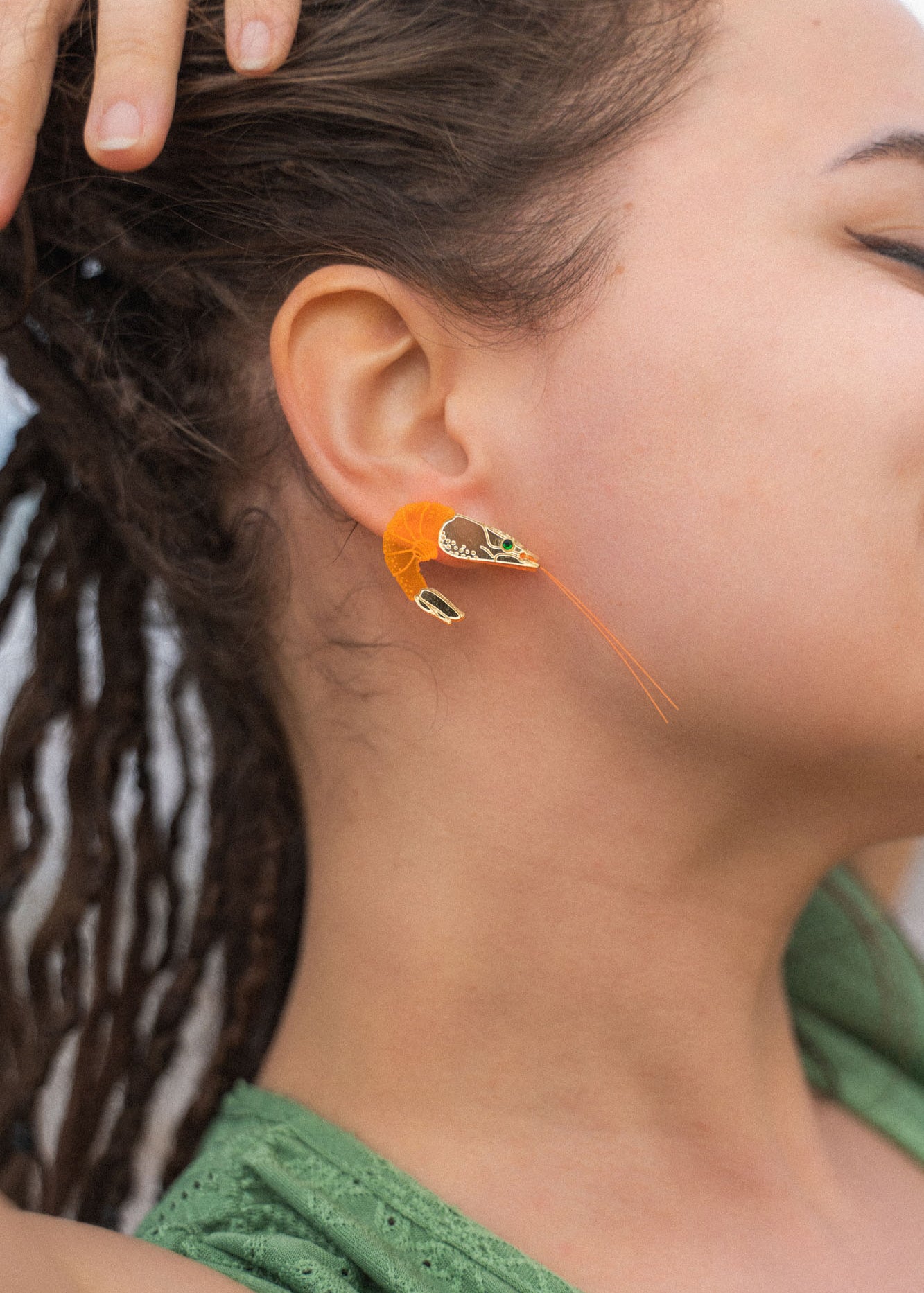 A person with brown skin and dreadlocks wears a detailed, neon orange Shrimp Stud earring adorned with emerald rhinestones. Their hand rests on their head, and they are dressed in a green top.