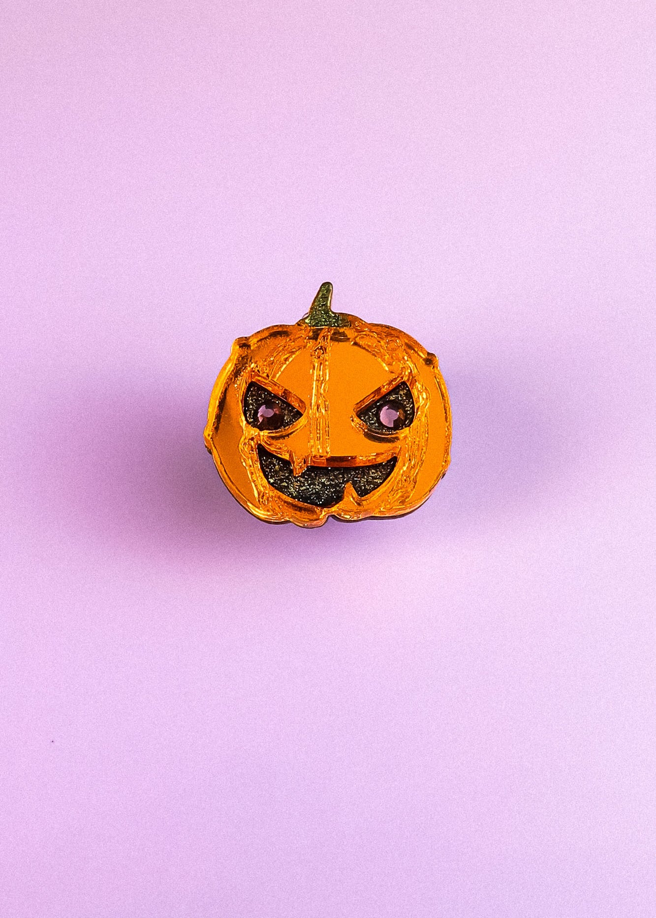 A small, shiny, orange Spooky Pumpkin Pin with a green stem and a mischievous expression sits on a plain light purple background, adding handcrafted Halloween charm to any décor.