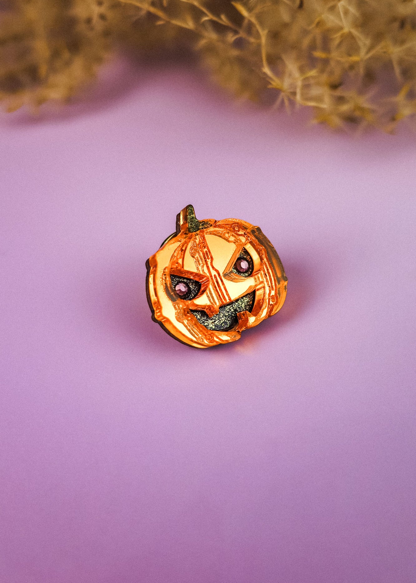 A small, handcrafted Spooky Pumpkin Pin with a mischievous face sits on a purple surface, perfect for adding a touch of Halloween charm amid blurred dried plant decorations in the background.