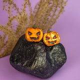 Two small spooky pumpkin studs with carved faces sit on a black rock, with dried grass in the background and a purple surface underneath—perfect Halloween earrings for fans of handcrafted jewelry.