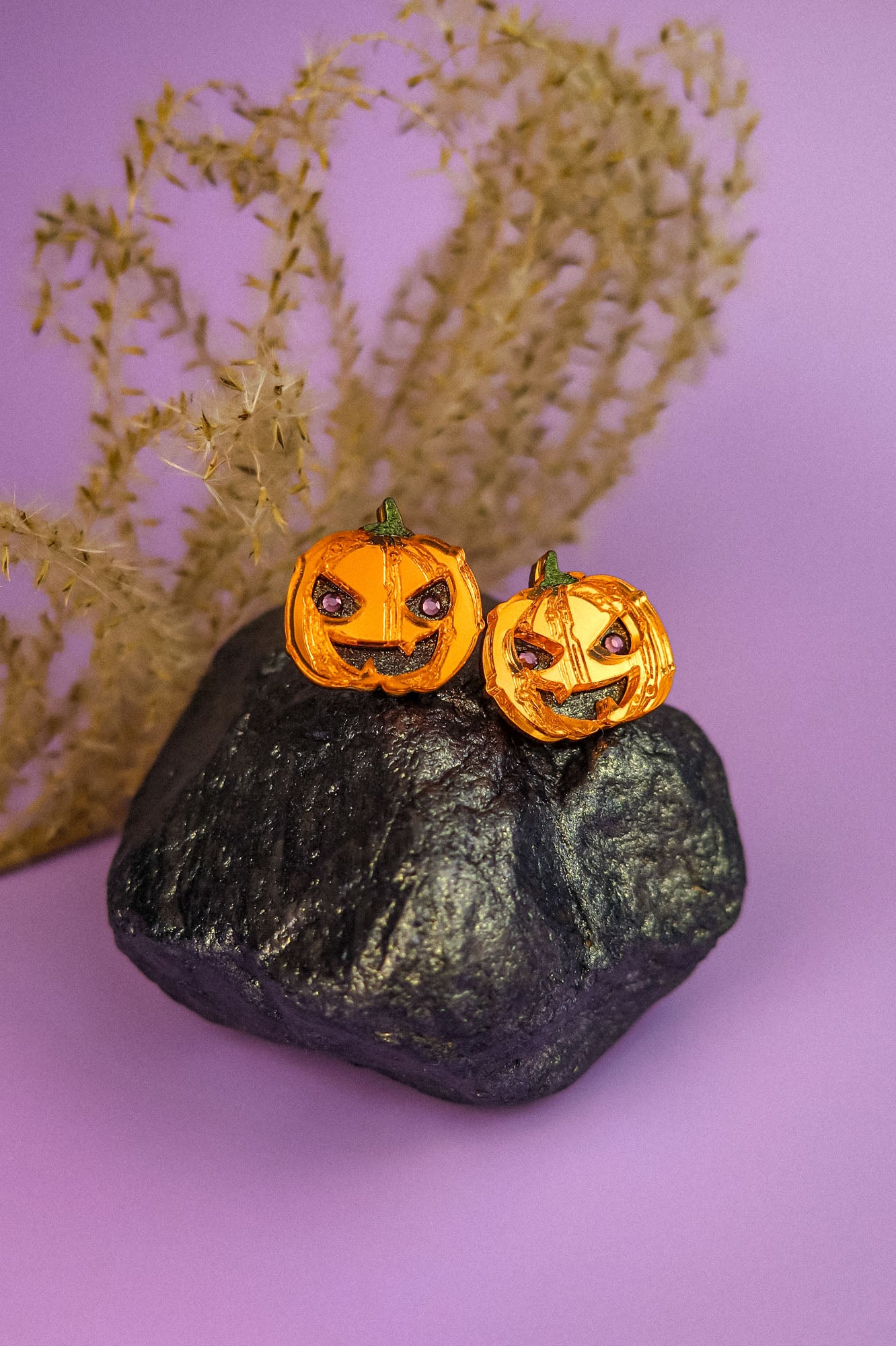 Two small spooky pumpkin studs with carved faces sit on a black rock, with dried grass in the background and a purple surface underneath—perfect Halloween earrings for fans of handcrafted jewelry.