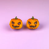 Two small jack-o-lanterns with angry expressions are placed side by side on a pink and purple gradient background, capturing the perfect mood for Spooky Pumpkin Studs—ideal handcrafted jewelry for Halloween earrings lovers.