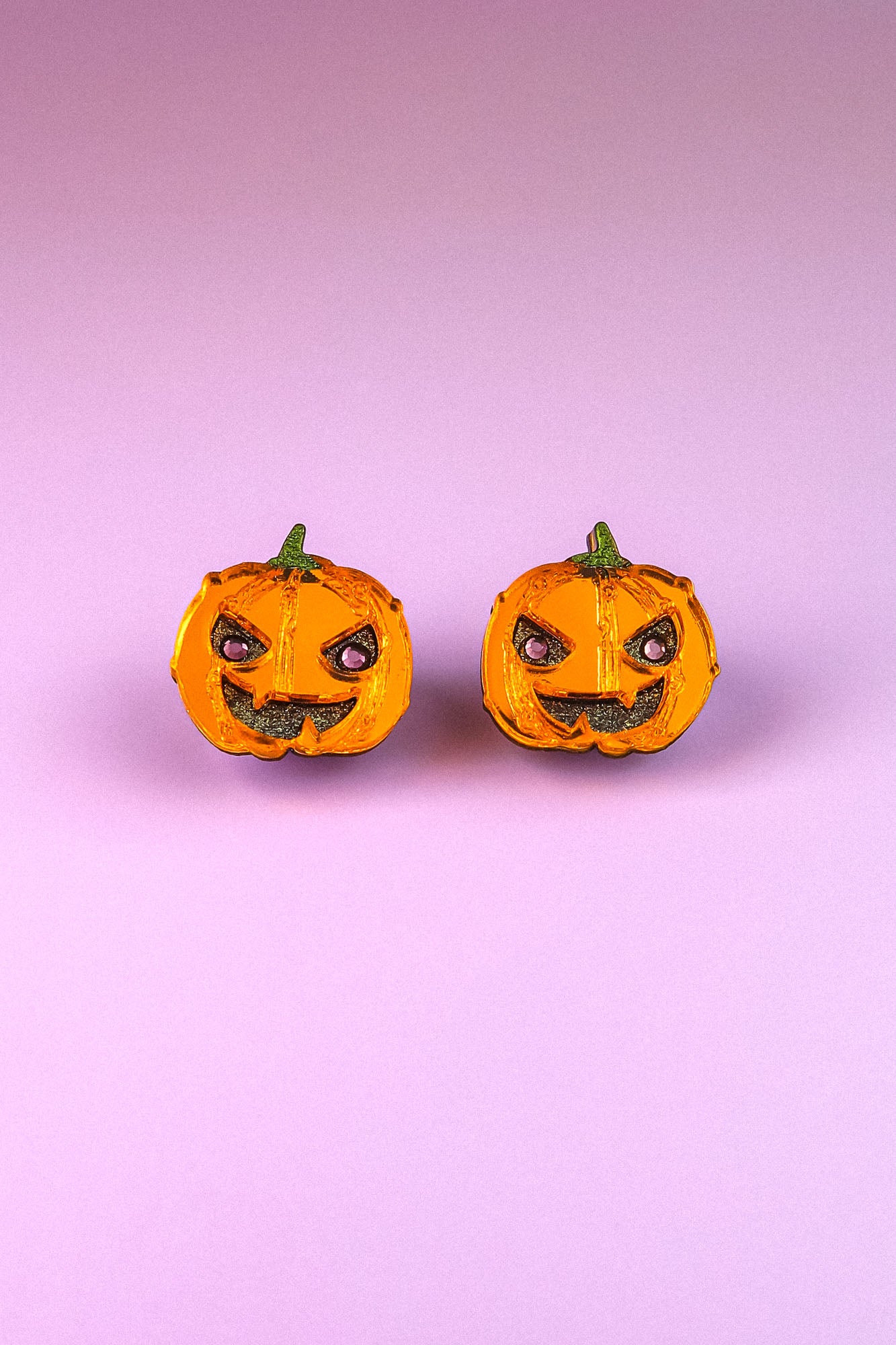 Two small jack-o-lanterns with angry expressions are placed side by side on a pink and purple gradient background, capturing the perfect mood for Spooky Pumpkin Studs—ideal handcrafted jewelry for Halloween earrings lovers.