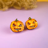 Two small, orange jack-o’-lantern decorations with mischievous expressions sit on a light purple surface, evoking the charm of Spooky Pumpkin Studs—perfect Halloween earrings for lovers of handcrafted jewelry. A dried plant rests in the yellow background.
