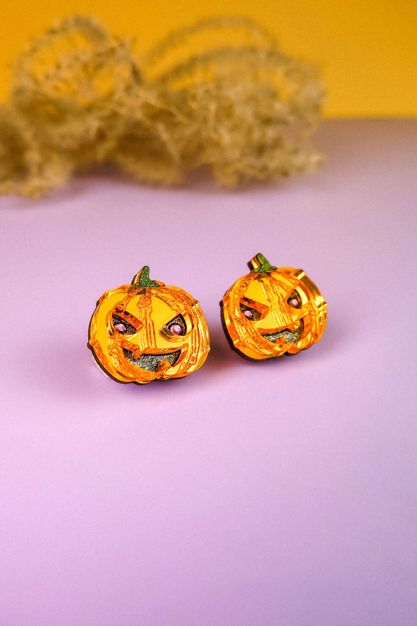 Two small, orange jack-o’-lantern decorations with mischievous expressions sit on a light purple surface, evoking the charm of Spooky Pumpkin Studs—perfect Halloween earrings for lovers of handcrafted jewelry. A dried plant rests in the yellow background.