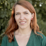 A woman with light skin and long red hair, wearing a green top and tiny heart studs, stands outdoors in front of green foliage, smiling softly at the camera.