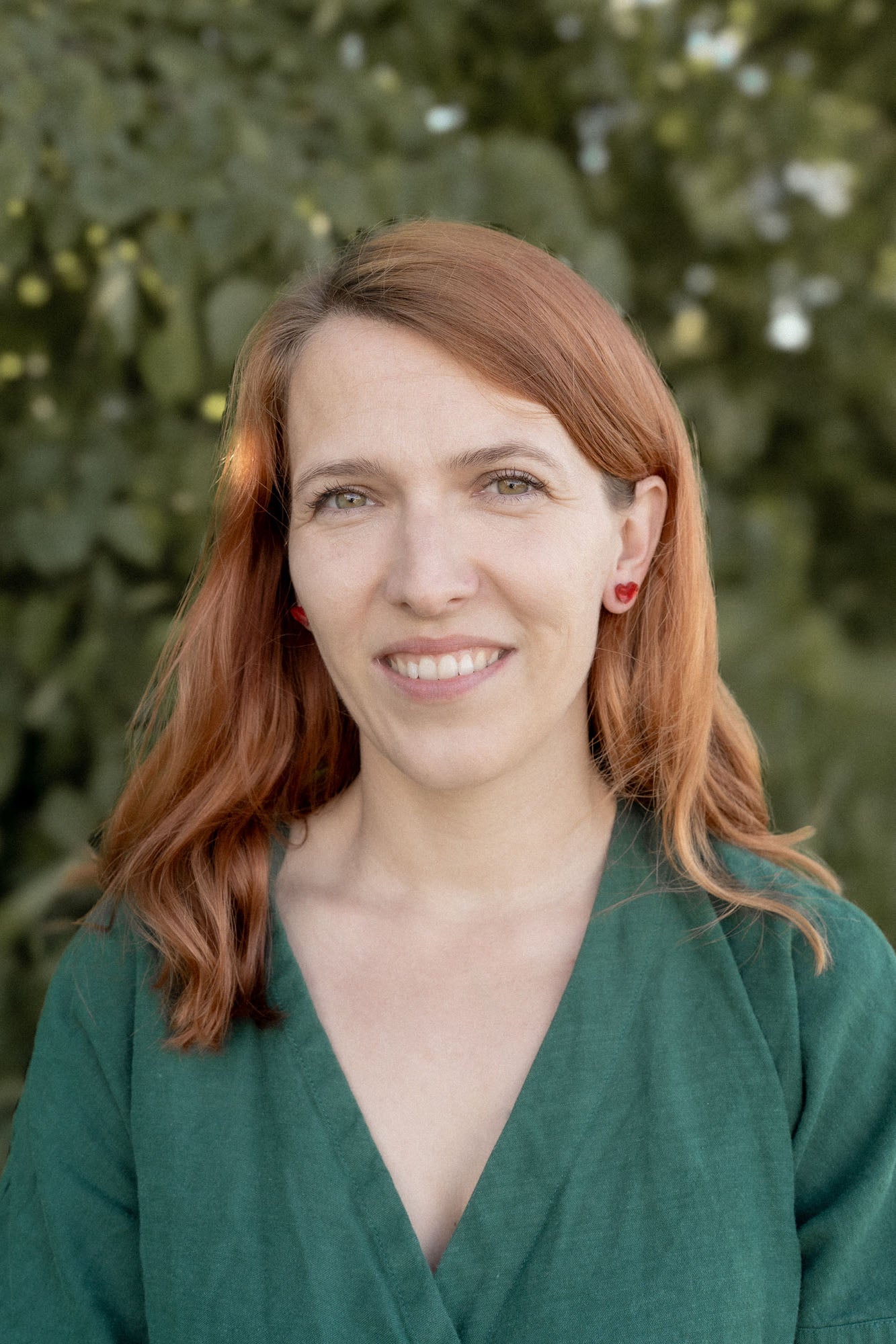 A woman with light skin and long red hair, wearing a green top and tiny heart studs, stands outdoors in front of green foliage, smiling softly at the camera.