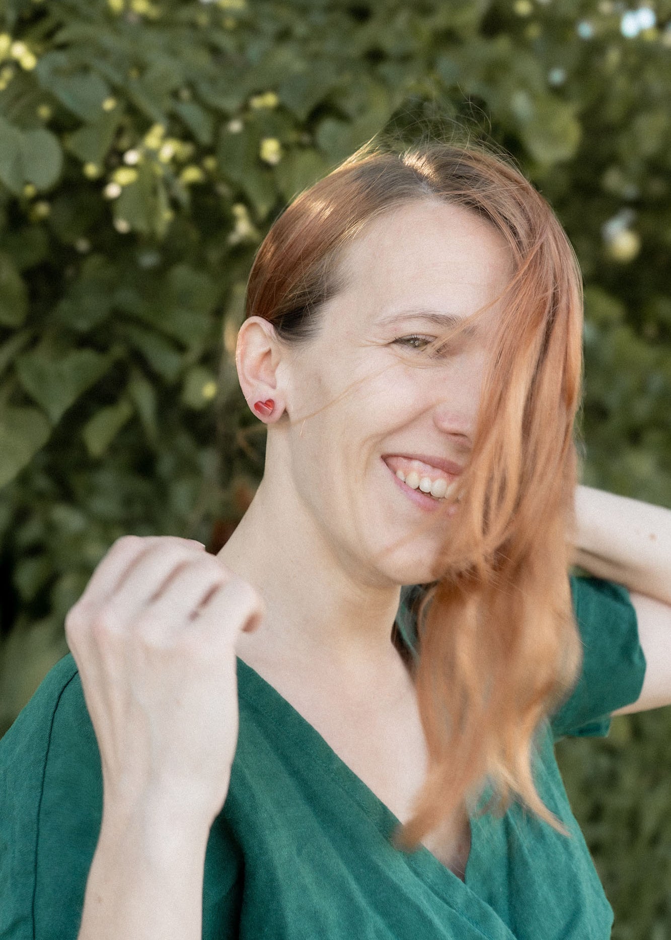 A woman with long, light brown hair wearing a green top smiles outdoors, tiny heart studs adding minimalist charm. She stands in front of leafy green foliage as sunlight filters through the trees, with some hair falling across her face.
