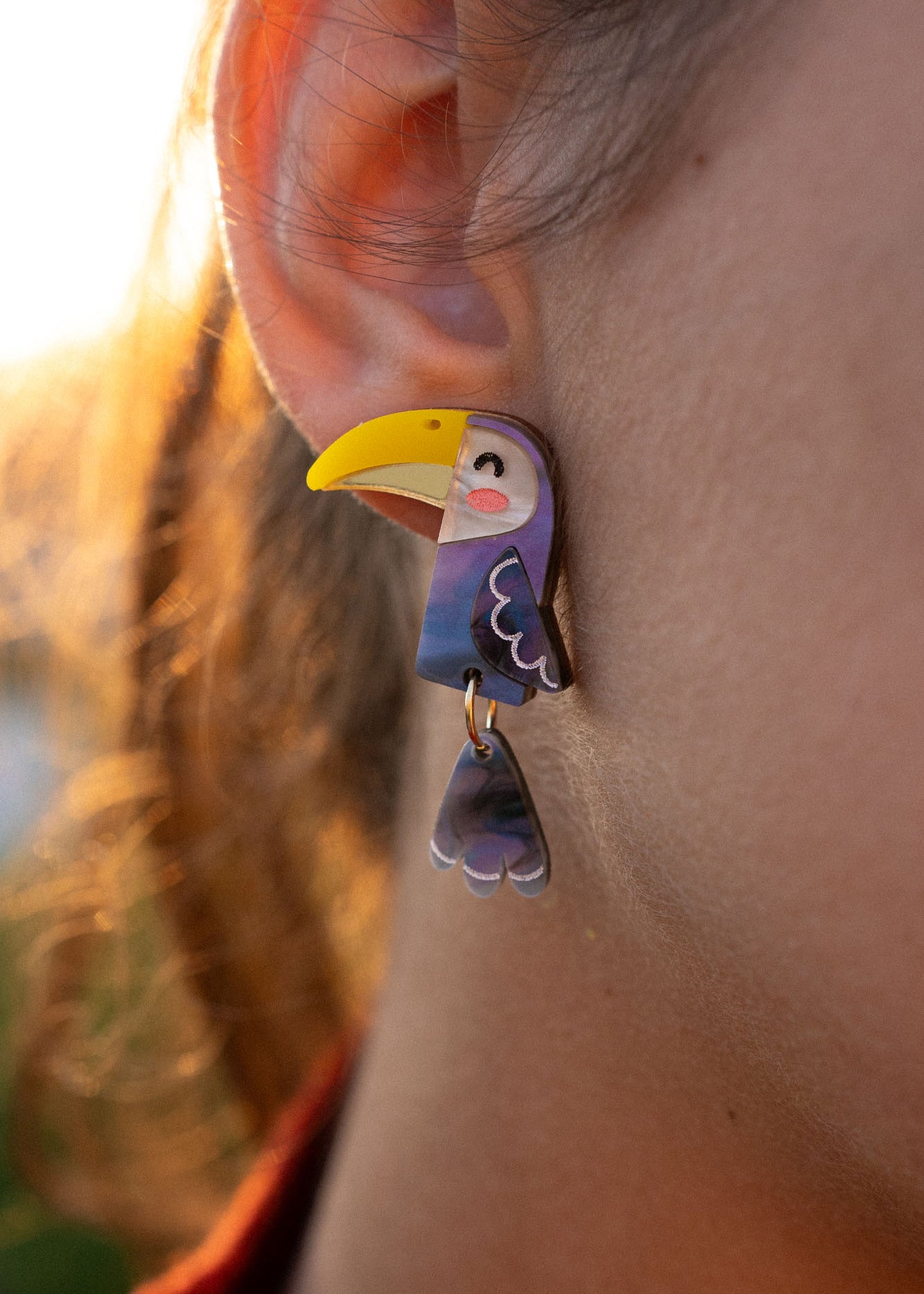 Close-up of a person’s ear adorned with handcrafted Toucan Earrings, featuring a whimsical yellow beak and dangling wing—a playful pop of color against the softly lit outdoor background.