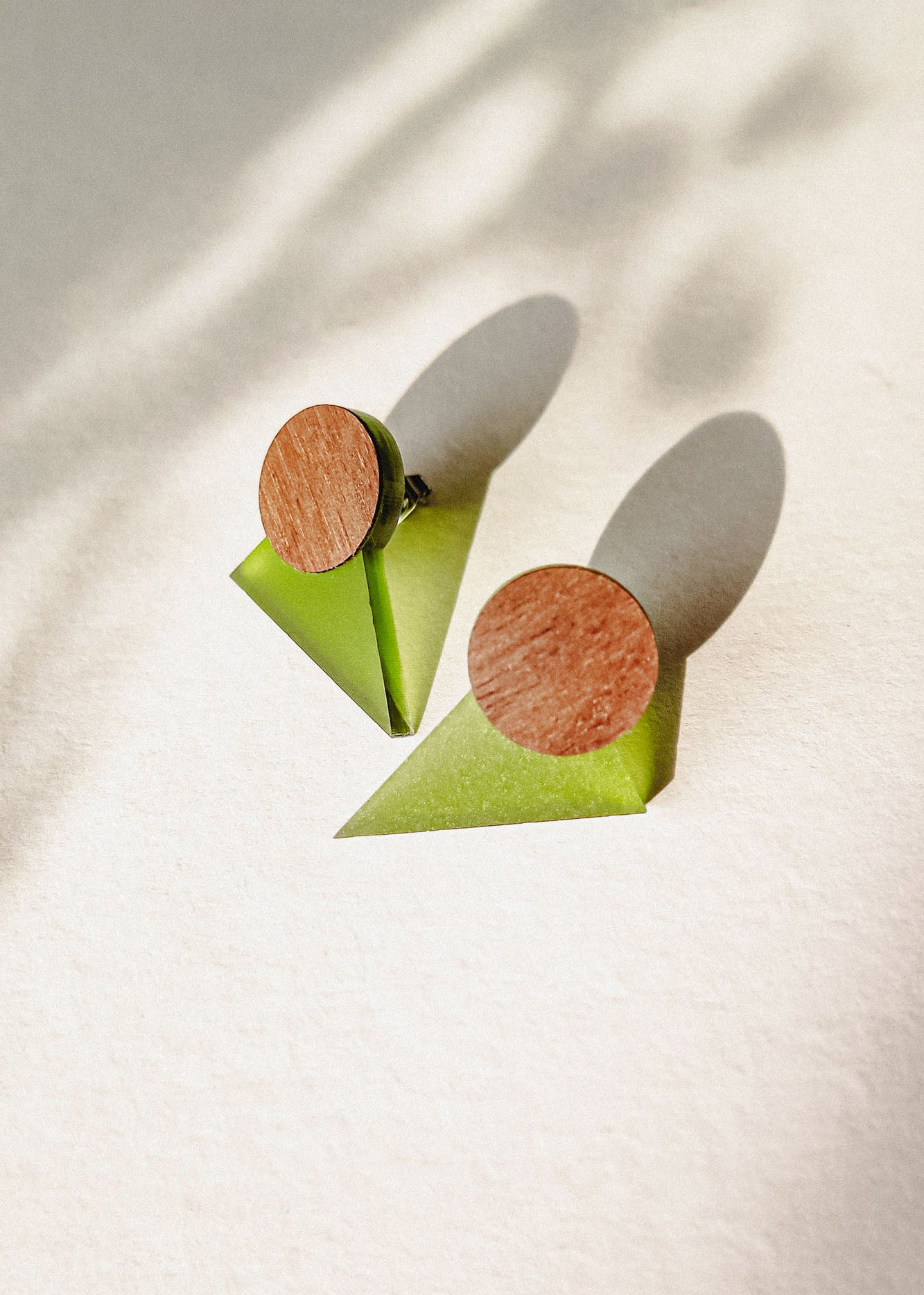 A pair of minimalist triangle earrings with round walnut wood tops and green acrylic glass bases are displayed on a light textured surface, casting soft shadows.