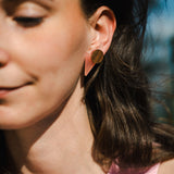 A woman with long brown hair wears striking triangle earrings made of acrylic glass, featuring pink and gold shapes. She is dressed in a light pink top and stands outdoors in sunlight, only the left side of her face visible with a soft smile.