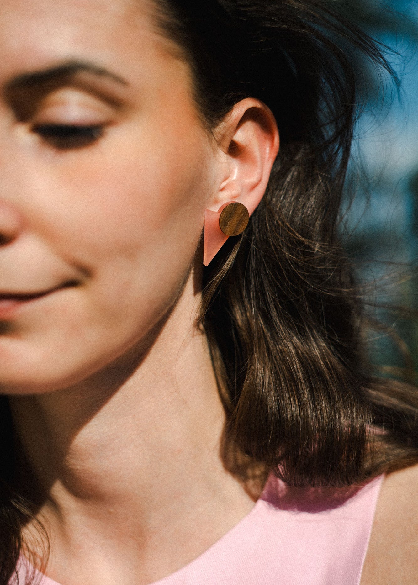 A woman with long brown hair wears striking triangle earrings made of acrylic glass, featuring pink and gold shapes. She is dressed in a light pink top and stands outdoors in sunlight, only the left side of her face visible with a soft smile.