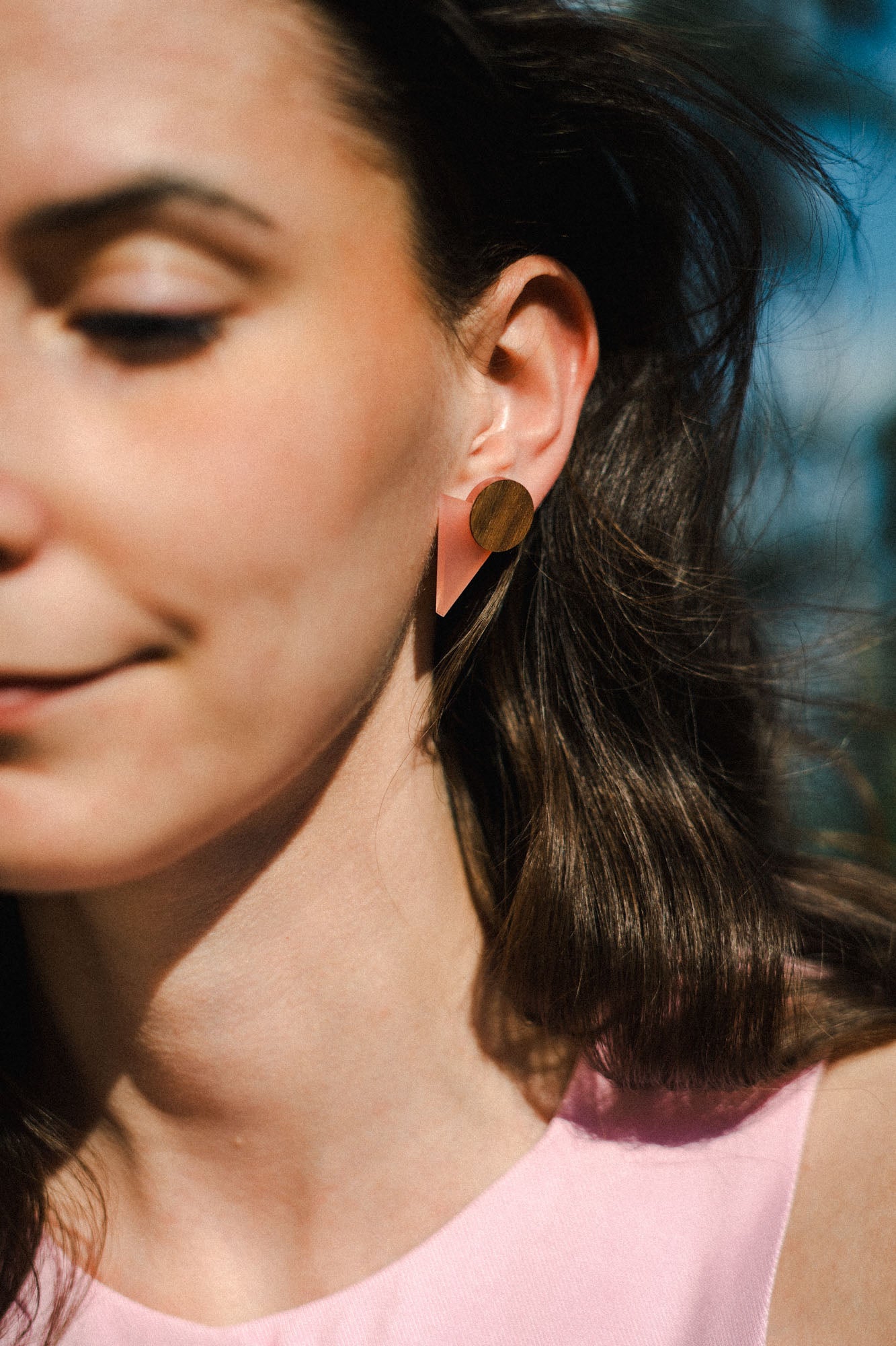 A woman with long brown hair wears striking triangle earrings made of acrylic glass, featuring pink and gold shapes. She is dressed in a light pink top and stands outdoors in sunlight, only the left side of her face visible with a soft smile.