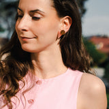 A woman with long brown hair, wearing a sleeveless pink top with pink buttons and chic triangle earrings, stands outdoors in sunlight, looking down and smiling softly. Trees and buildings are blurred in the background.