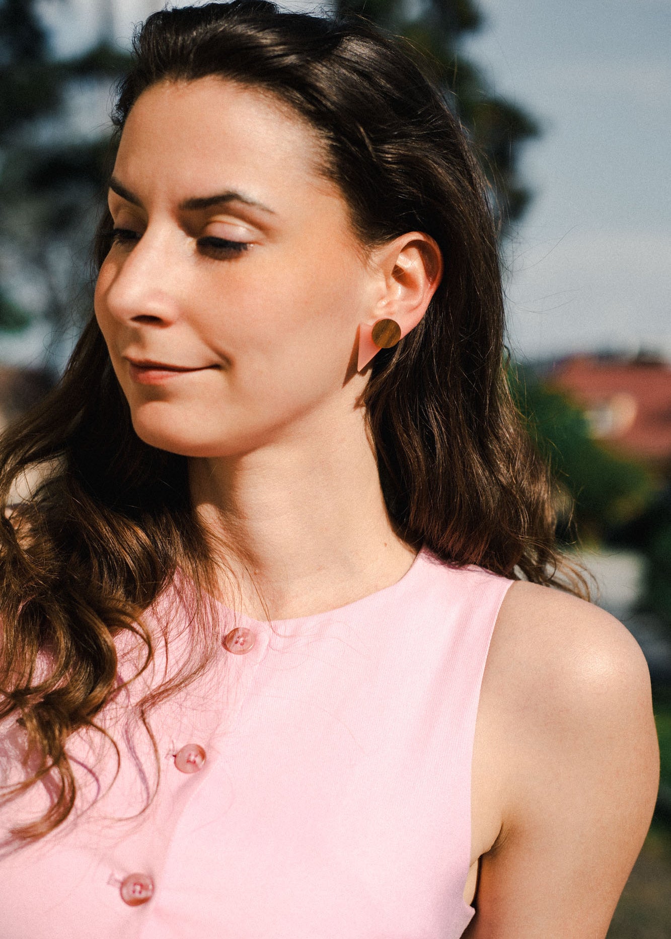 A woman with long brown hair, wearing a sleeveless pink top with pink buttons and chic triangle earrings, stands outdoors in sunlight, looking down and smiling softly. Trees and buildings are blurred in the background.