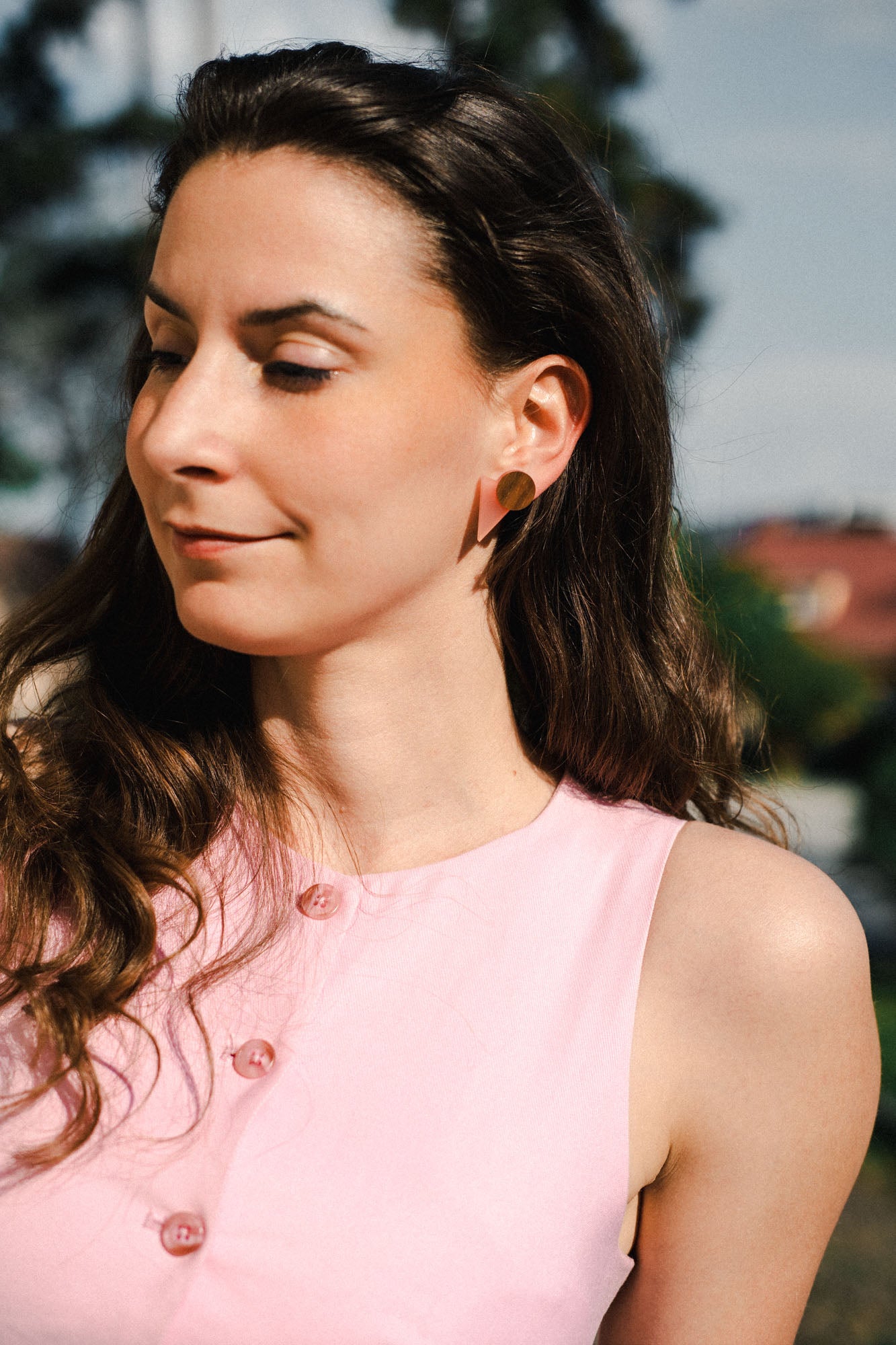 A woman with long brown hair, wearing a sleeveless pink top with pink buttons and chic triangle earrings, stands outdoors in sunlight, looking down and smiling softly. Trees and buildings are blurred in the background.