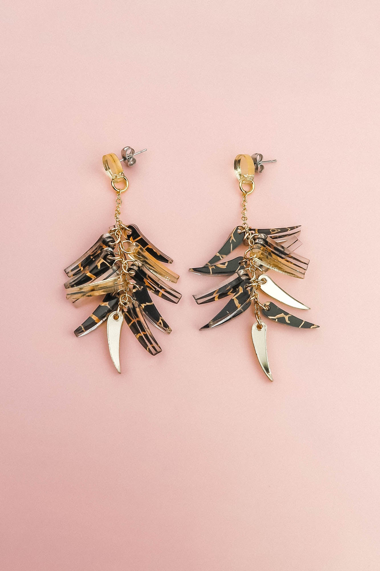 A pair of featherlight dangle earrings with layered gold and tortoiseshell-patterned pieces arranged in a fan-like design, displayed on a light pink background.