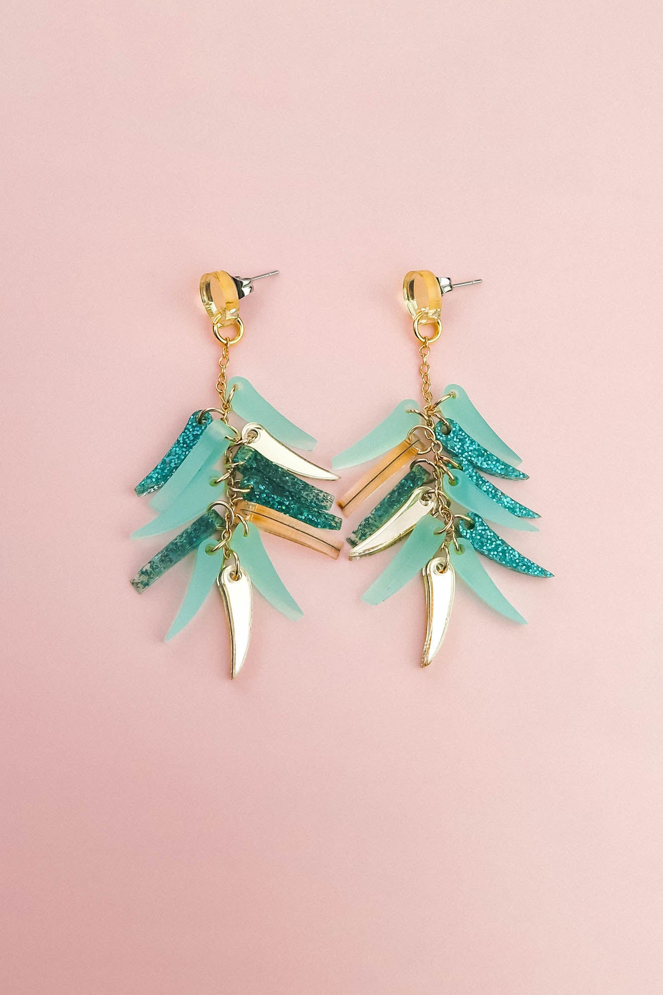 A pair of dangling earrings with gold hardware, featuring layered teal, blue glitter, and gold leaf-shaped charms, these unique style earrings are displayed on a light pink background.