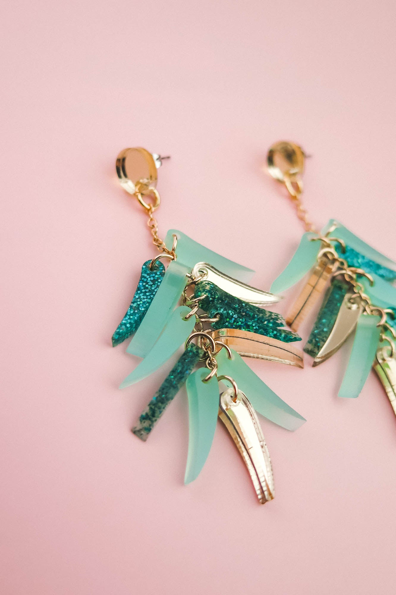 A pair of gold dangle earrings with teal and glittery blue acrylic accents, these unique style earrings are displayed on a pale pink background.