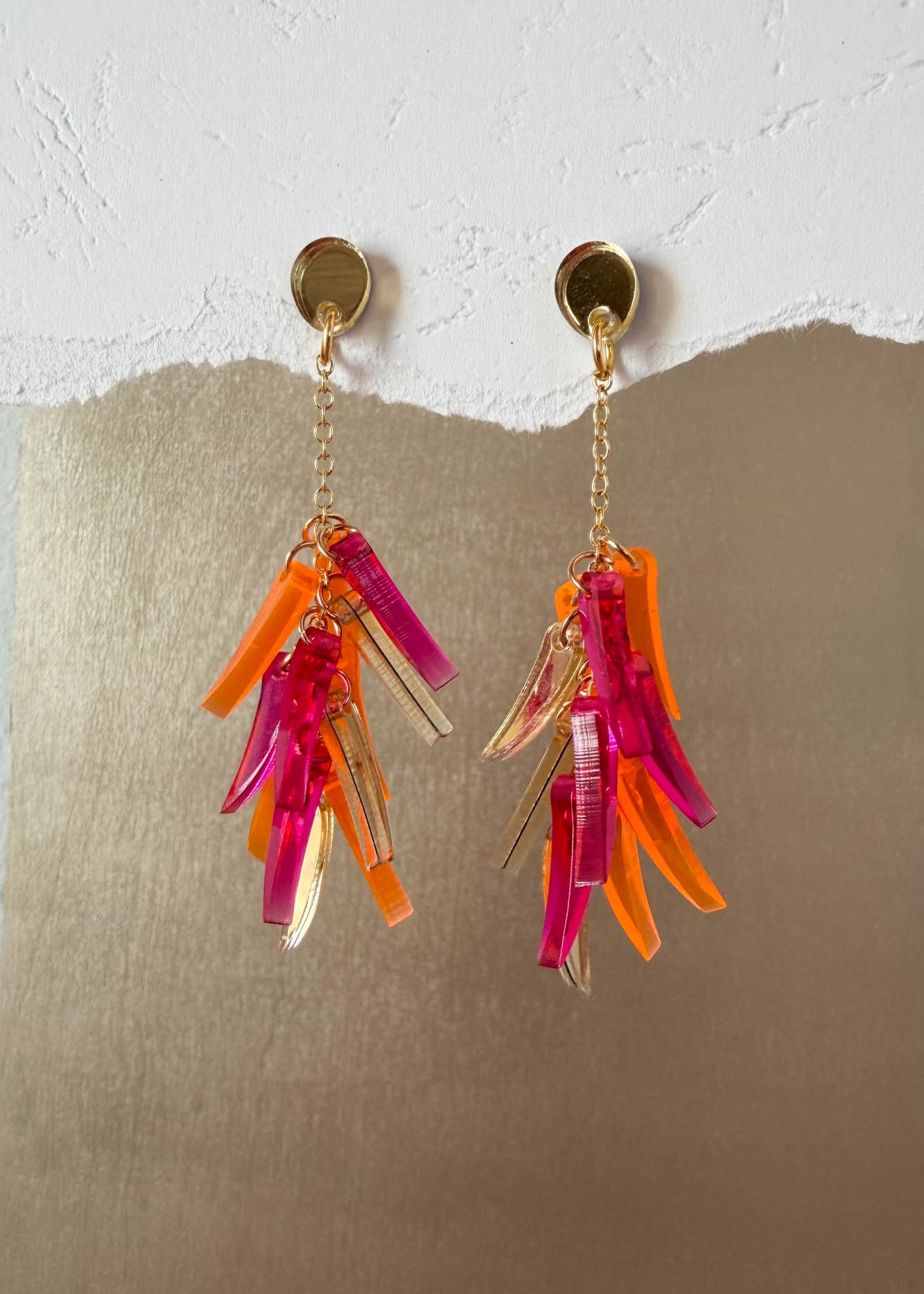 A pair of featherlight dangle earrings with gold studs and chains, each featuring clusters of translucent pink, orange, and clear acrylic shards, displayed against a textured white and metallic gold background.