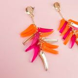 A pair of featherlight dangle earrings on a pink background, featuring gold metal accents and colorful acrylic charms in orange, pink, and gold.