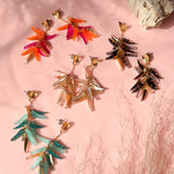 Colorful featherlight dangle earrings in four pairs—red, orange, teal, and black—are arranged on a pink surface with a rock and delicate dried grass, all showcased in natural sunlight.