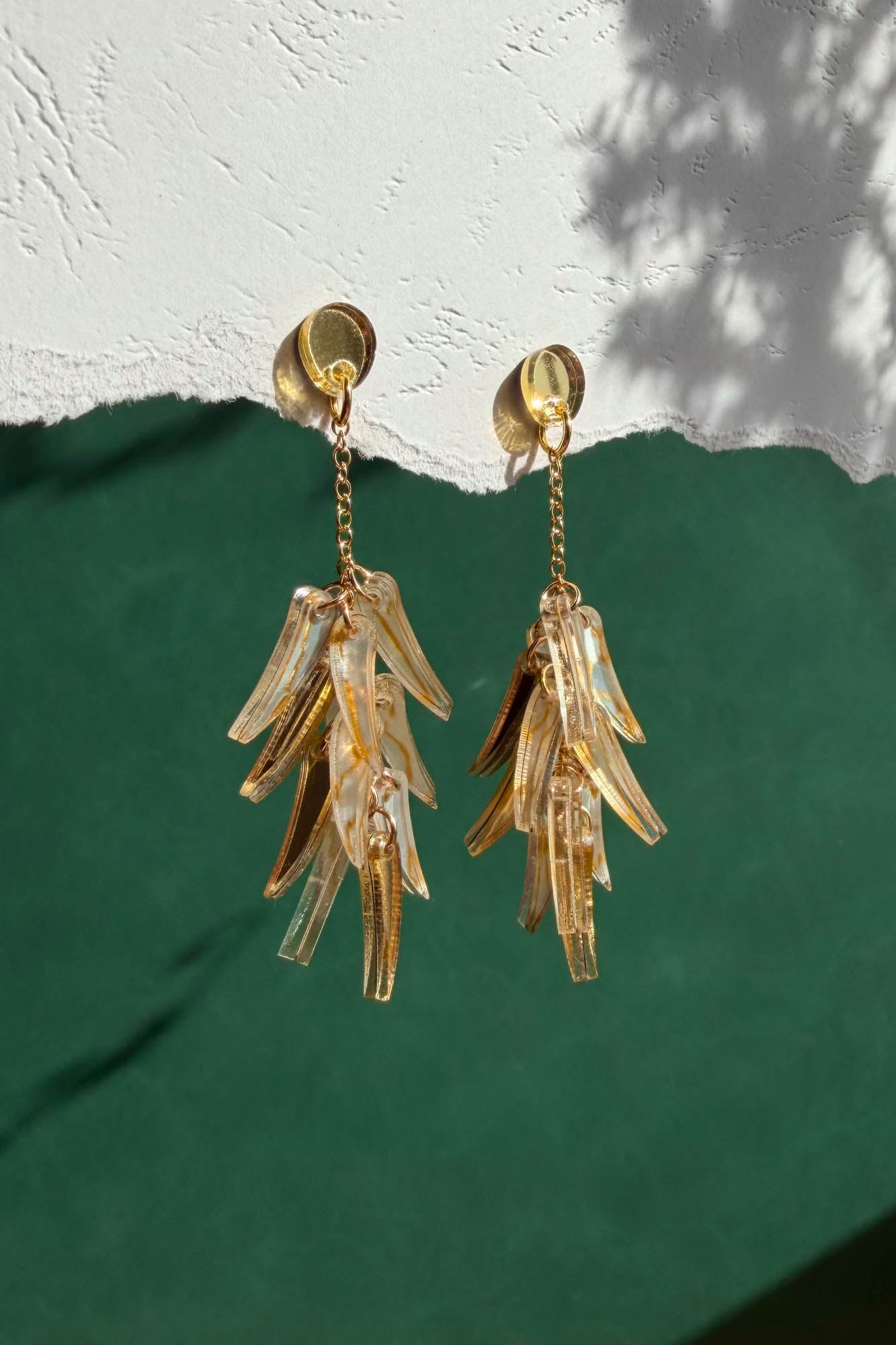 A pair of featherlight, gold chain dangle earrings with translucent, leaf-shaped pendants hang against a textured white and green background with shadow patterns.