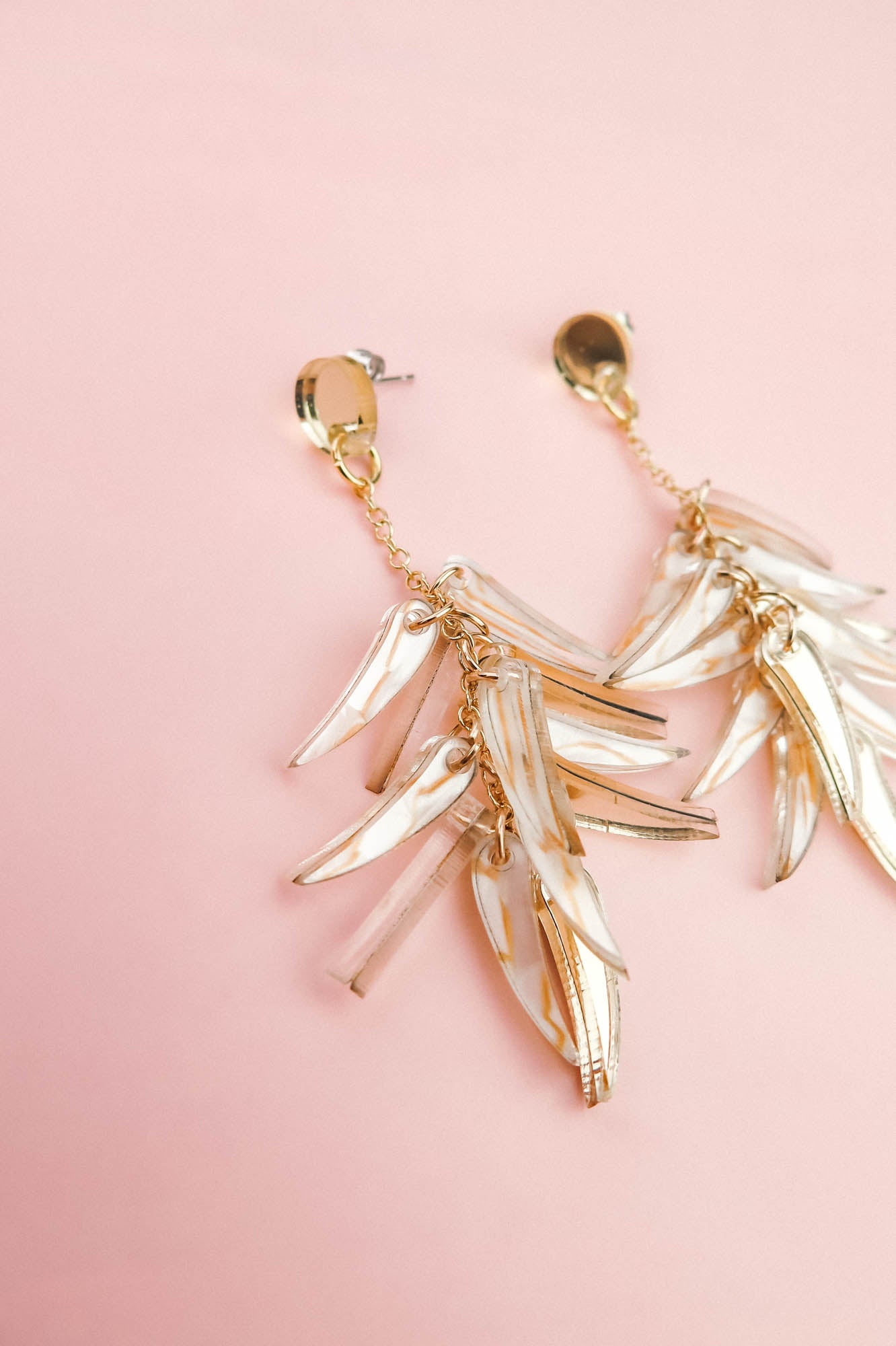 A pair of featherlight gold dangle earrings with leaf-shaped, translucent accents, displayed on a soft pink background.