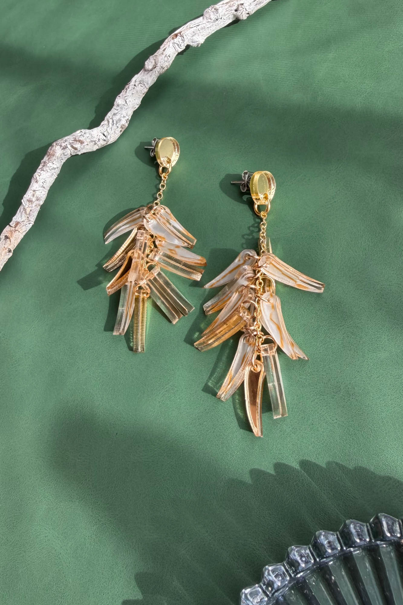 A pair of gold and clear acrylic dangle earrings shaped like layered leaves, these featherlight earrings are displayed on a green surface with textured driftwood and a partial glass dish.