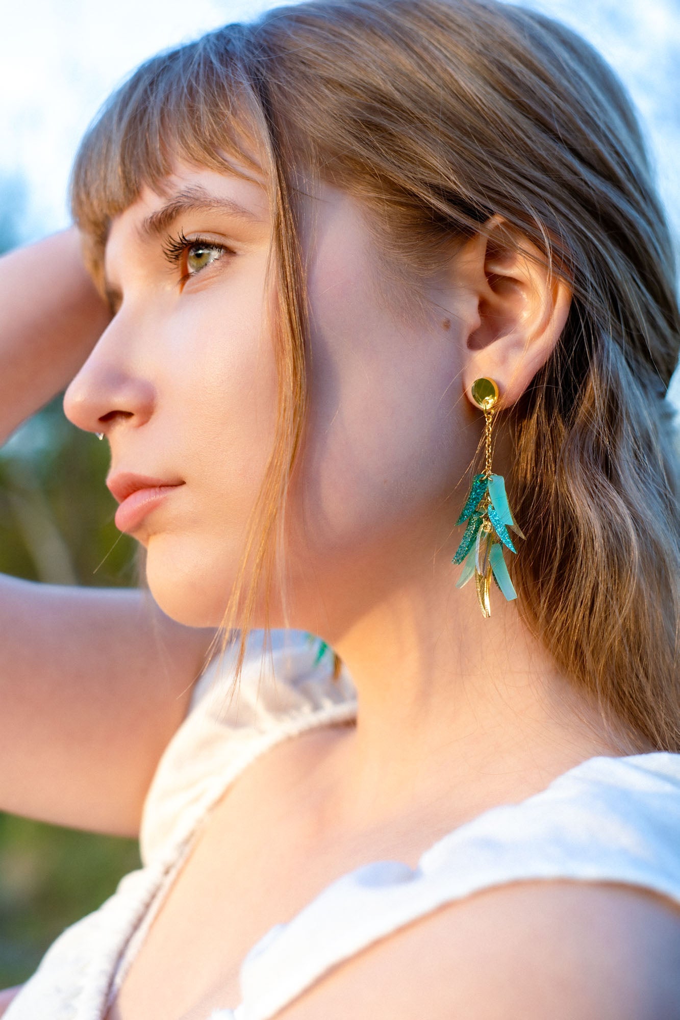 A young woman with light brown hair, wearing a white top and featherlight dangle earrings shaped like colorful feathers, gazes thoughtfully into the distance in soft natural light.