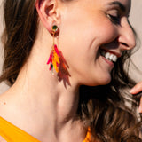 A woman with long brown hair smiles while wearing a sleeveless orange top and featherlight dangle earrings with pink, red, and yellow accents. The image highlights her earrings and natural makeup.