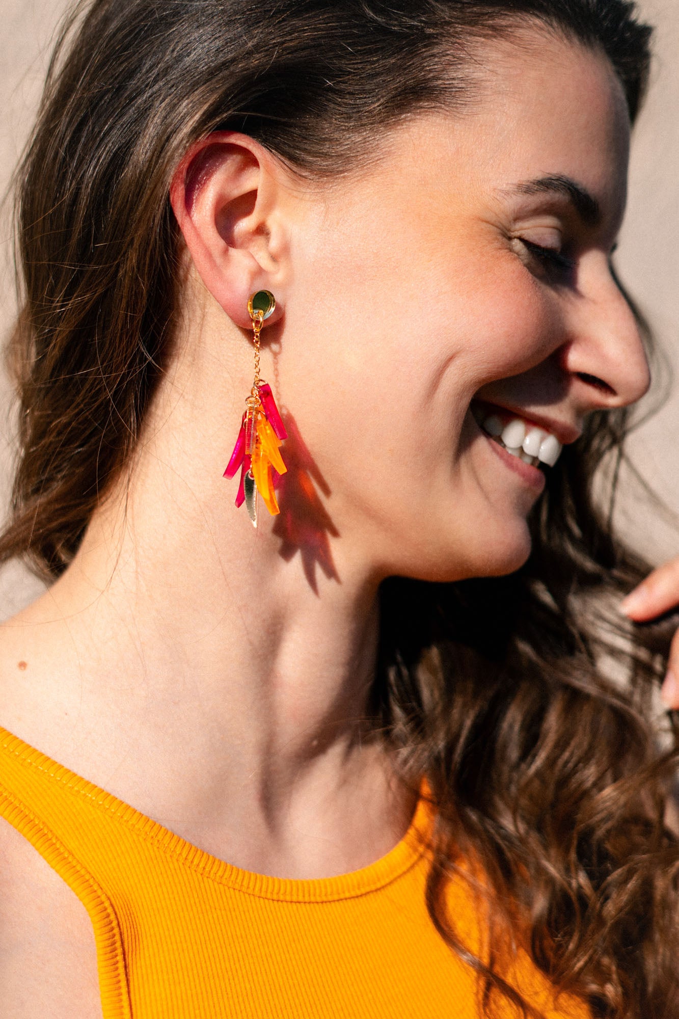 A woman with long brown hair smiles while wearing a sleeveless orange top and featherlight dangle earrings with pink, red, and yellow accents. The image highlights her earrings and natural makeup.