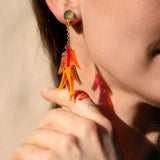 Close-up of a woman wearing featherlight dangle earrings shaped like orange flower petals. She is smiling, and her hand with a delicate tattoo touches her neck near the earring. Her hair is tucked behind her ear.