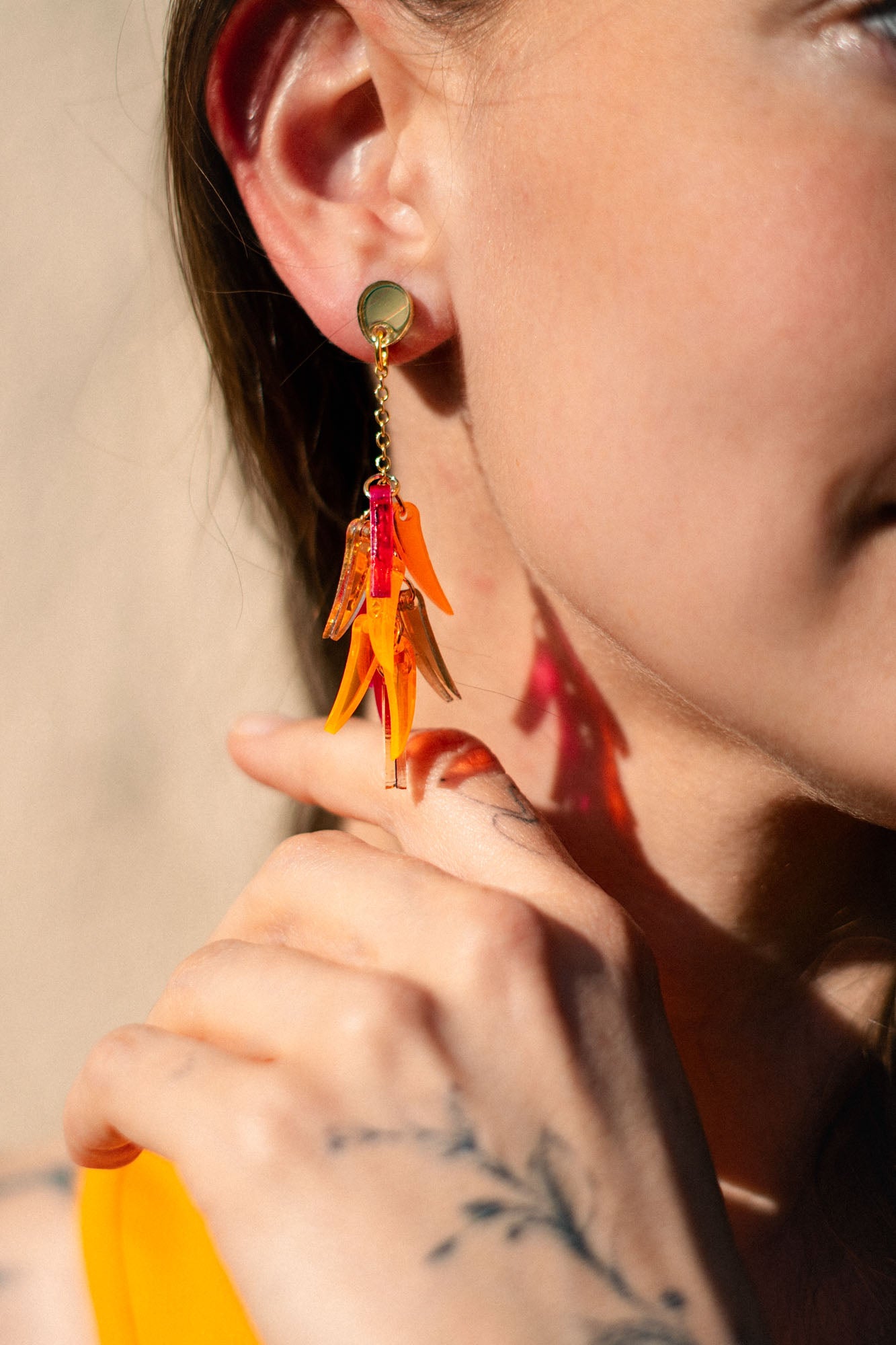 Close-up of a woman wearing featherlight dangle earrings shaped like orange flower petals. She is smiling, and her hand with a delicate tattoo touches her neck near the earring. Her hair is tucked behind her ear.