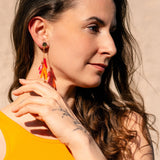 A woman with long brown hair wearing a sleeveless yellow top and featherlight acrylic tufts looks to the side, touching her earring with her hand. Sunlight highlights her face against a neutral background.
