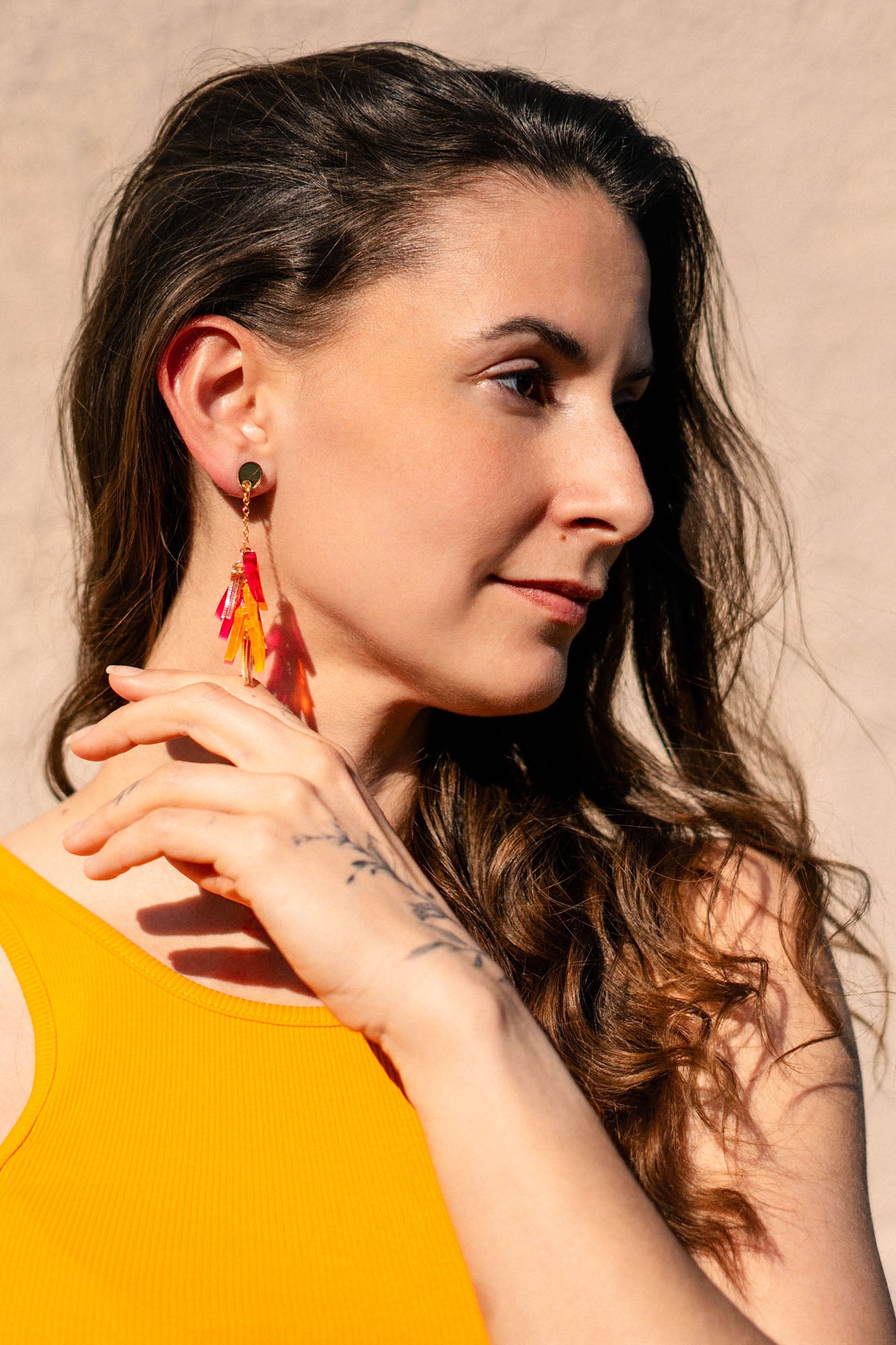 A woman with long brown hair wearing a sleeveless yellow top and featherlight acrylic tufts looks to the side, touching her earring with her hand. Sunlight highlights her face against a neutral background.