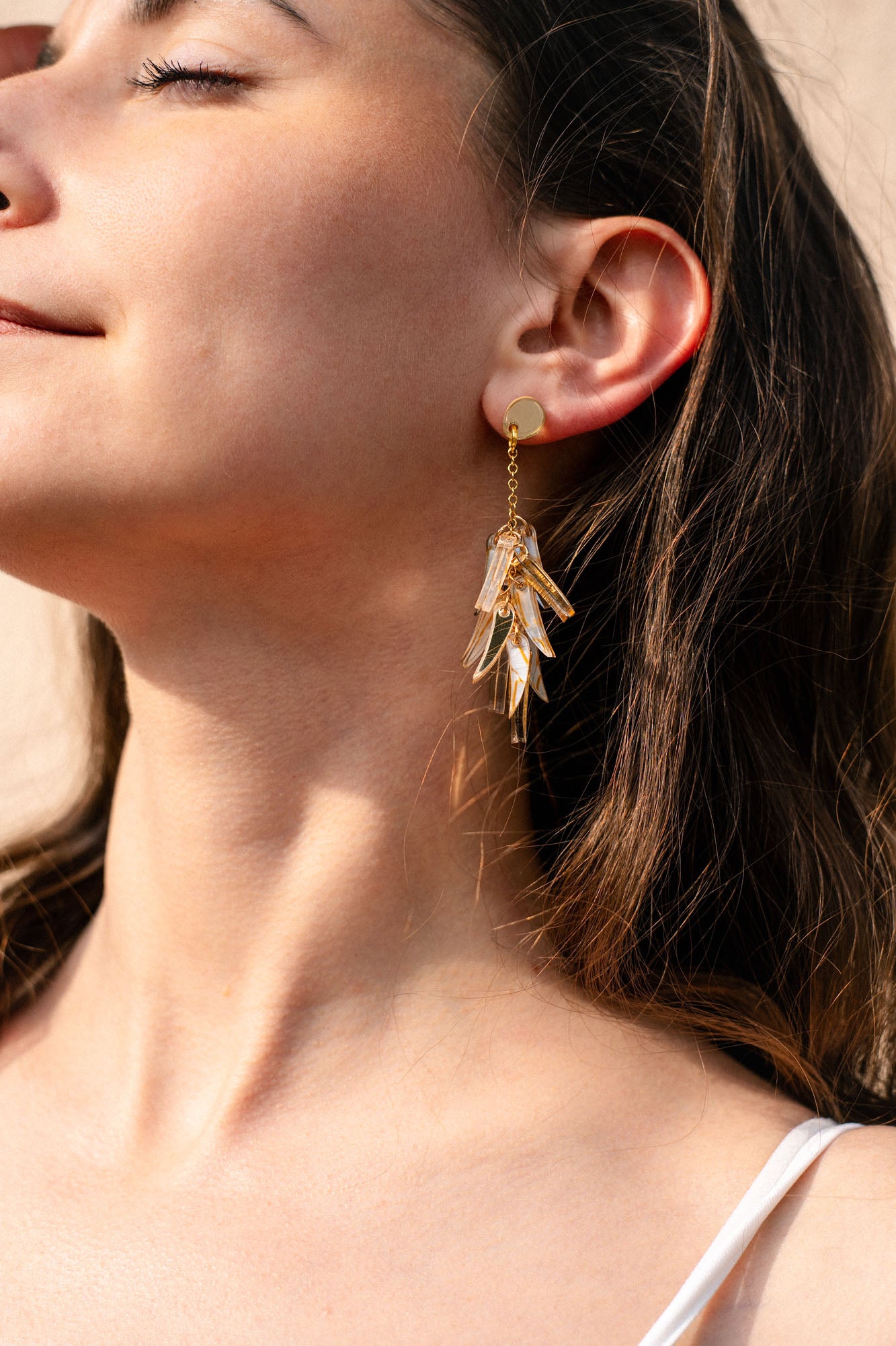 Close-up of a woman with long brown hair, eyes closed, and a slight smile, wearing featherlight gold dangle earrings shaped like cascading leaves. She is dressed in a white top as sunlight highlights her skin.