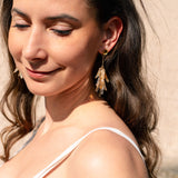 A woman with long brown hair and a white sleeveless top smiles gently with her eyes closed. She wears featherlight gold and crystal dangle earrings, standing in sunlight against a light-colored background.