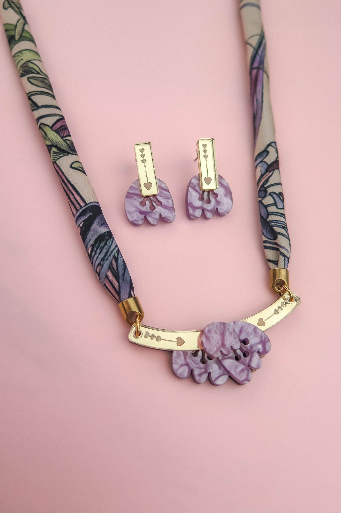 A patterned fabric handmade necklace with gold accents and a pendant featuring marbled purple shapes, paired with matching marbled purple earrings, is displayed on a soft pink background.