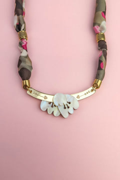 A handmade necklace with a gold curved bar, white shell-like pieces, and a multicolored fabric chain reminiscent of a silk-print scarf in green, pink, brown, and gold accents, displayed on a light pink background.