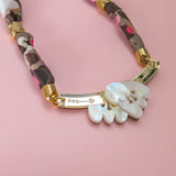 A close-up of a handmade necklace with a fabric-wrapped chain in brown, beige, and pink, gold details, and a white, carved mother-of-pearl pendant on a pink background.