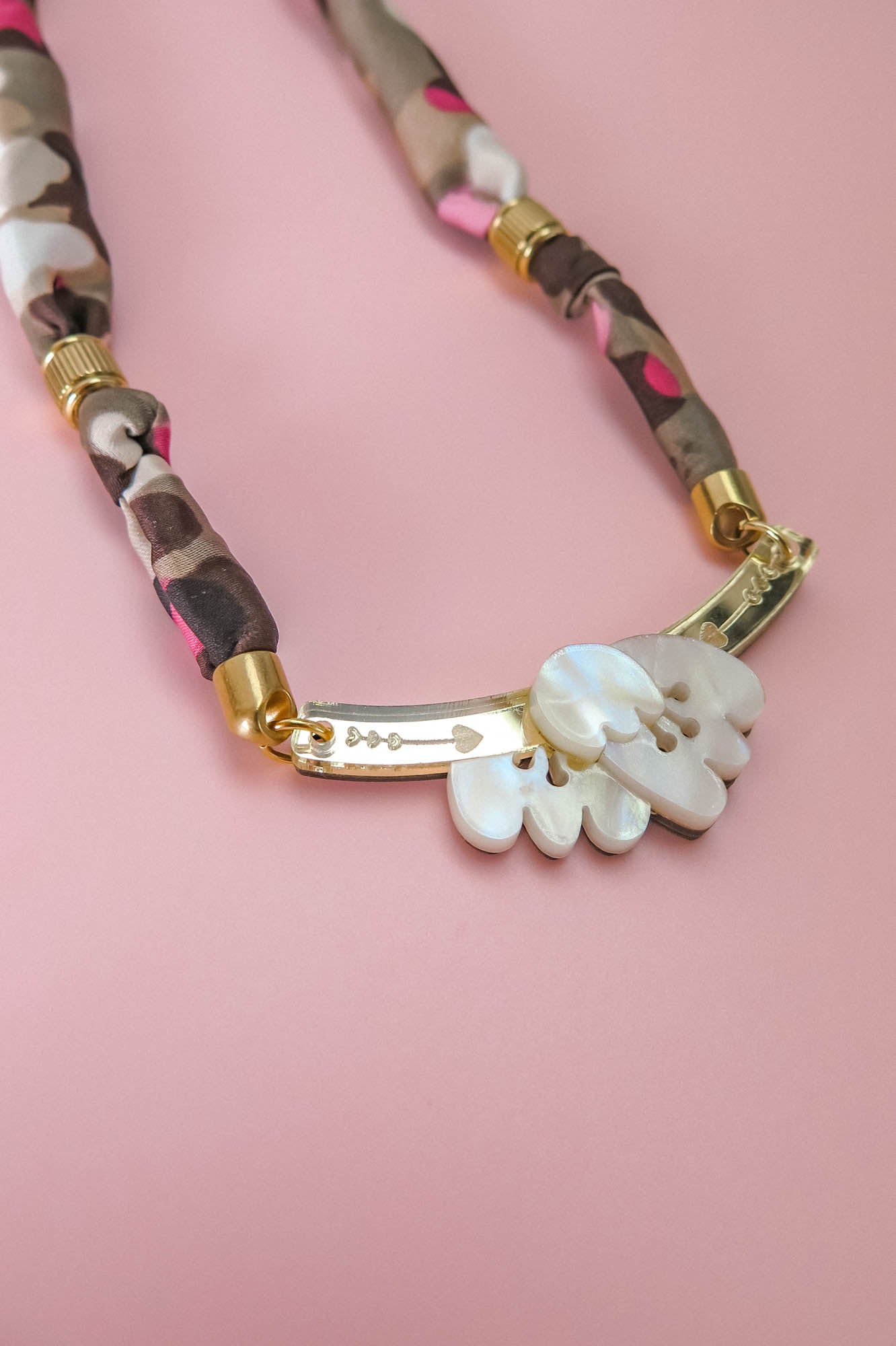 A close-up of a handmade necklace with a fabric-wrapped chain in brown, beige, and pink, gold details, and a white, carved mother-of-pearl pendant on a pink background.