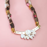 A handmade necklace with a pink, brown, and beige fabric band, gold accents, and a curved gold bar adorned with five white shell-like charms is displayed on a pink background.