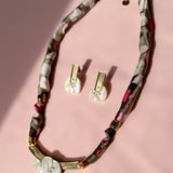 A handmade necklace and matching earrings are displayed on a pink surface. The necklace features a patterned fabric cord, gold accents, and a white floral pendant, while the earrings have white floral pieces with gold details and acrylic tulip touches.