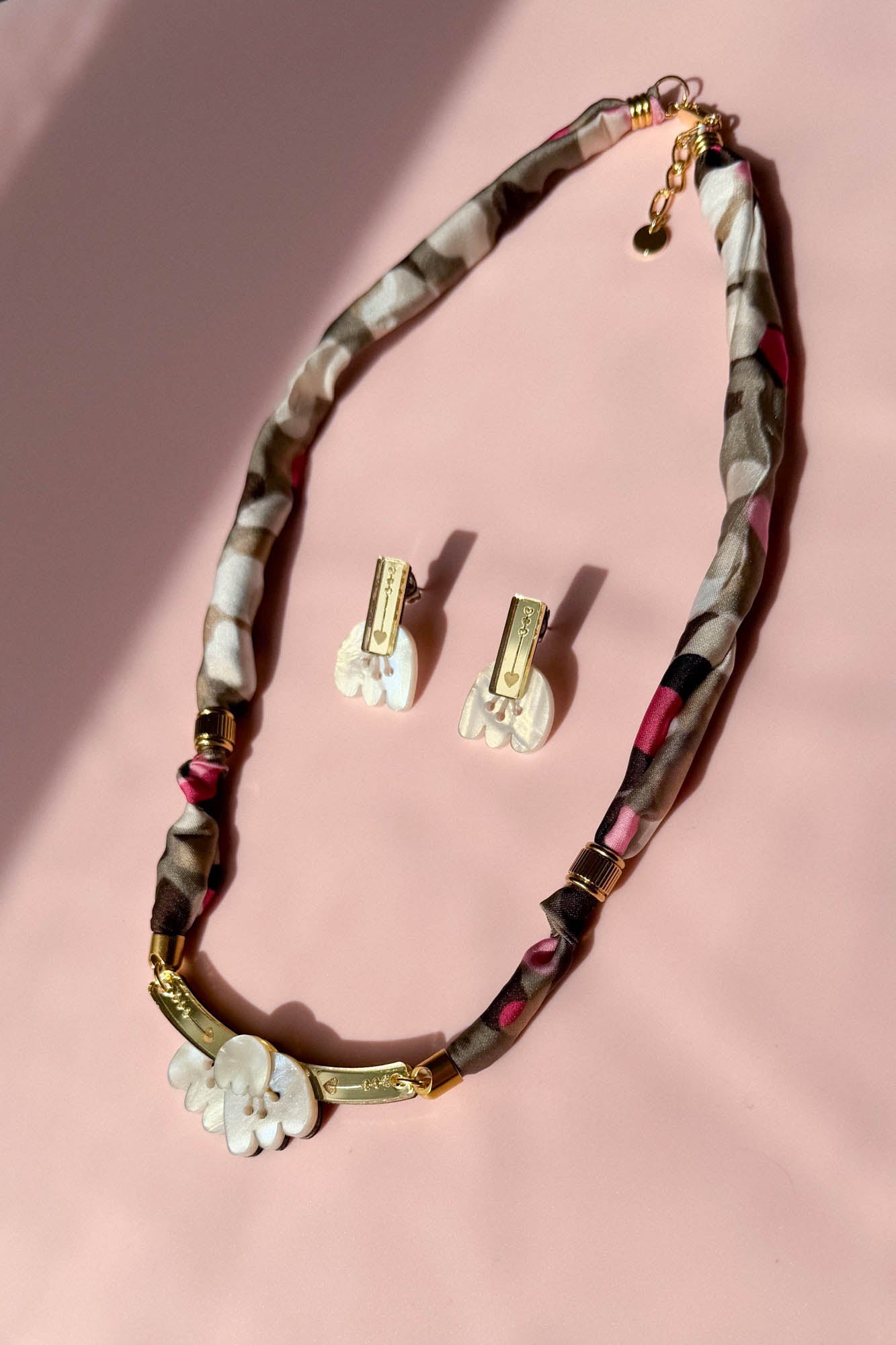A handmade necklace and matching earrings are displayed on a pink surface. The necklace features a patterned fabric cord, gold accents, and a white floral pendant, while the earrings have white floral pieces with gold details and acrylic tulip touches.