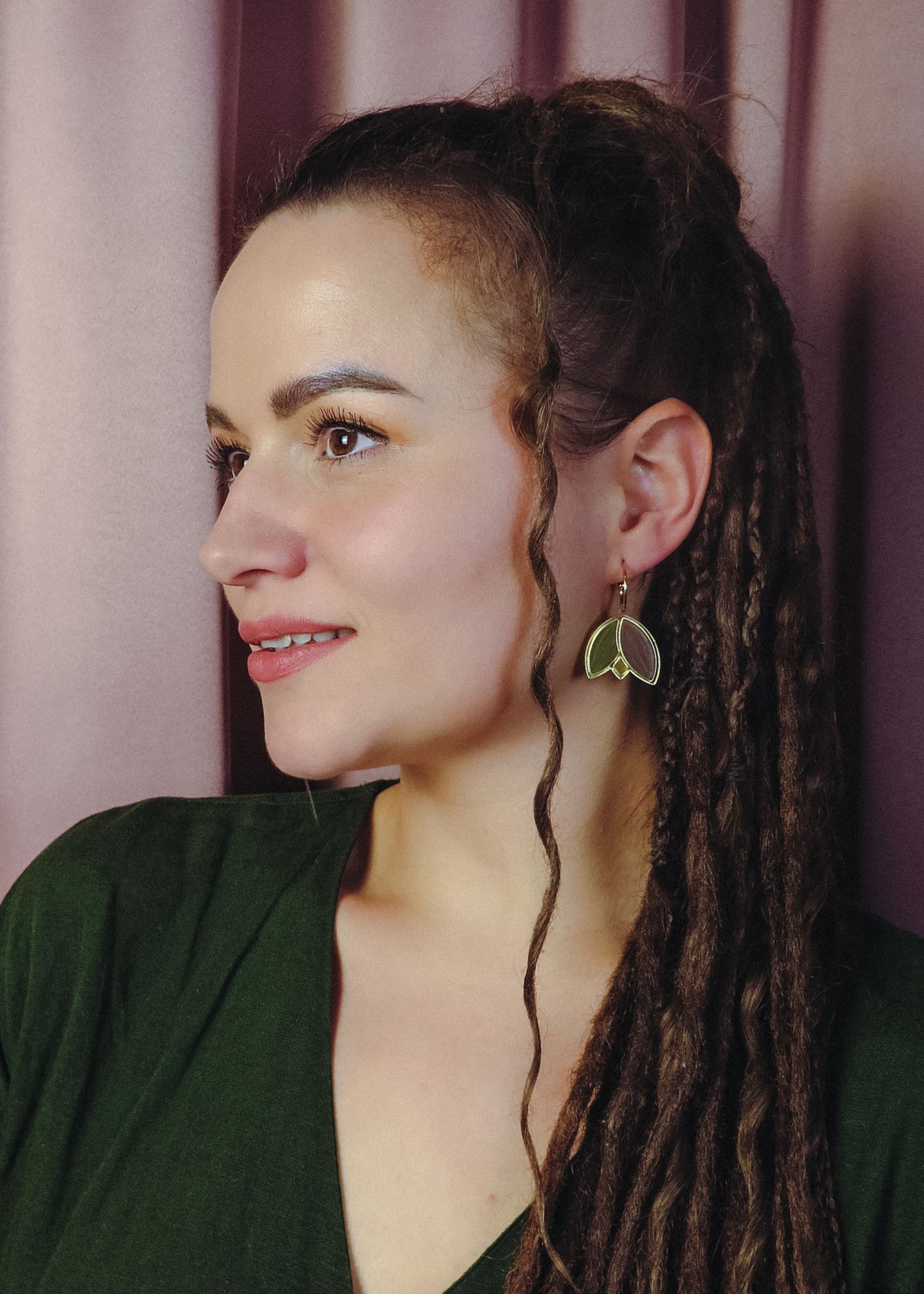 A woman with long, brown dreadlocks and a loose curl framing her face wears handcrafted leaf-shaped earrings and a green top, gazing to the left against a backdrop of soft pink curtains.