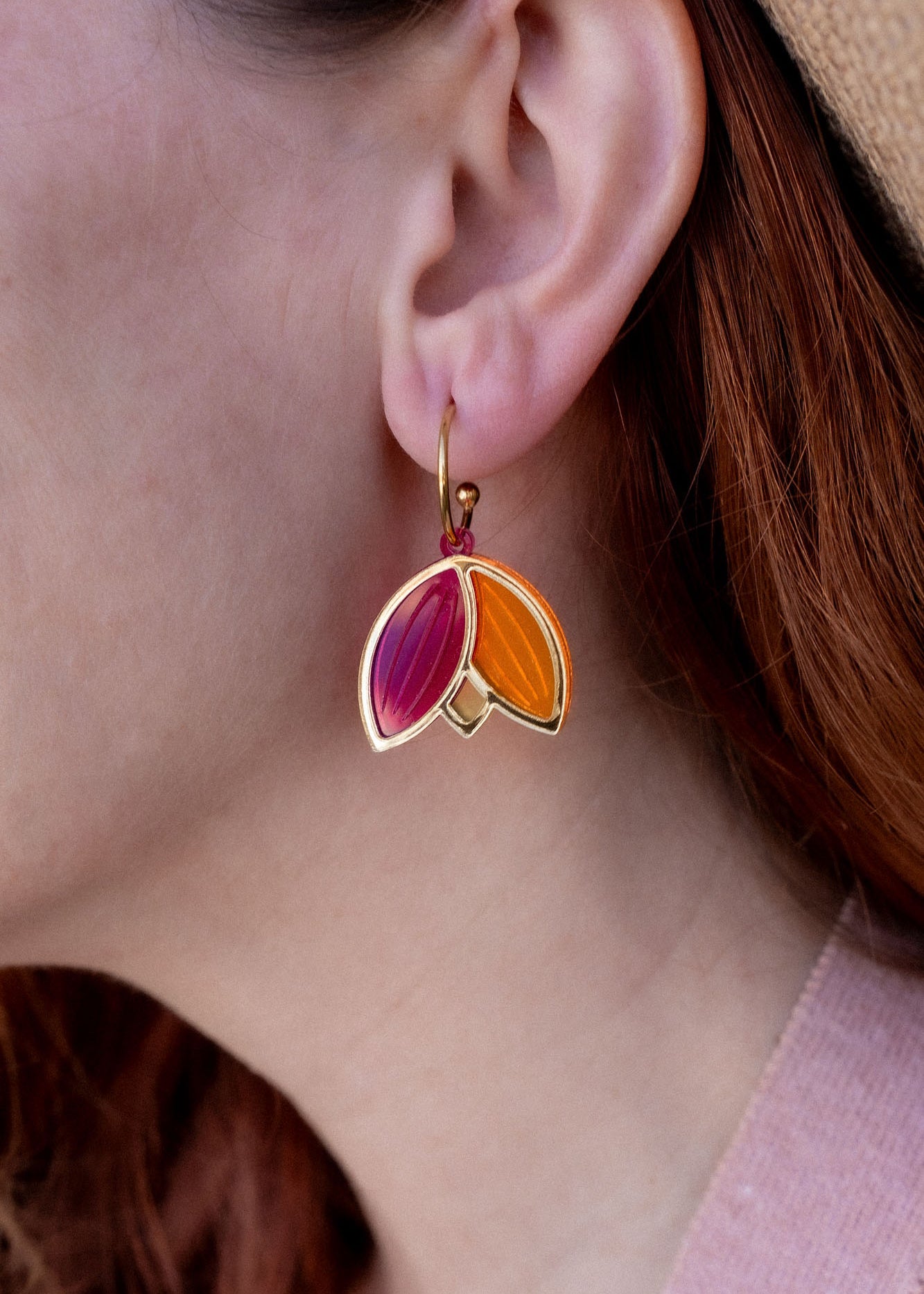 A person with red hair wearing a tan hat and pink sweater is shown in profile, displaying Tulip Huggie Earrings crafted with artisan craftsmanship and featuring gold, pink, and orange enamel sections.