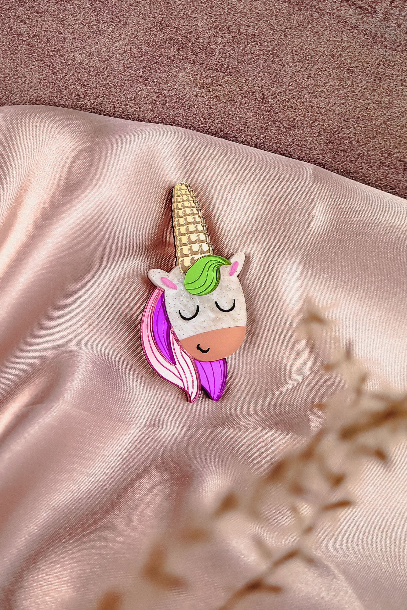 A handcrafted UniCorn Brooch with a gold horn, green and pink mane, and closed eyes rests on soft, shiny pink fabric. A blurred plant is visible in the foreground, highlighting this playful accessory’s charm.