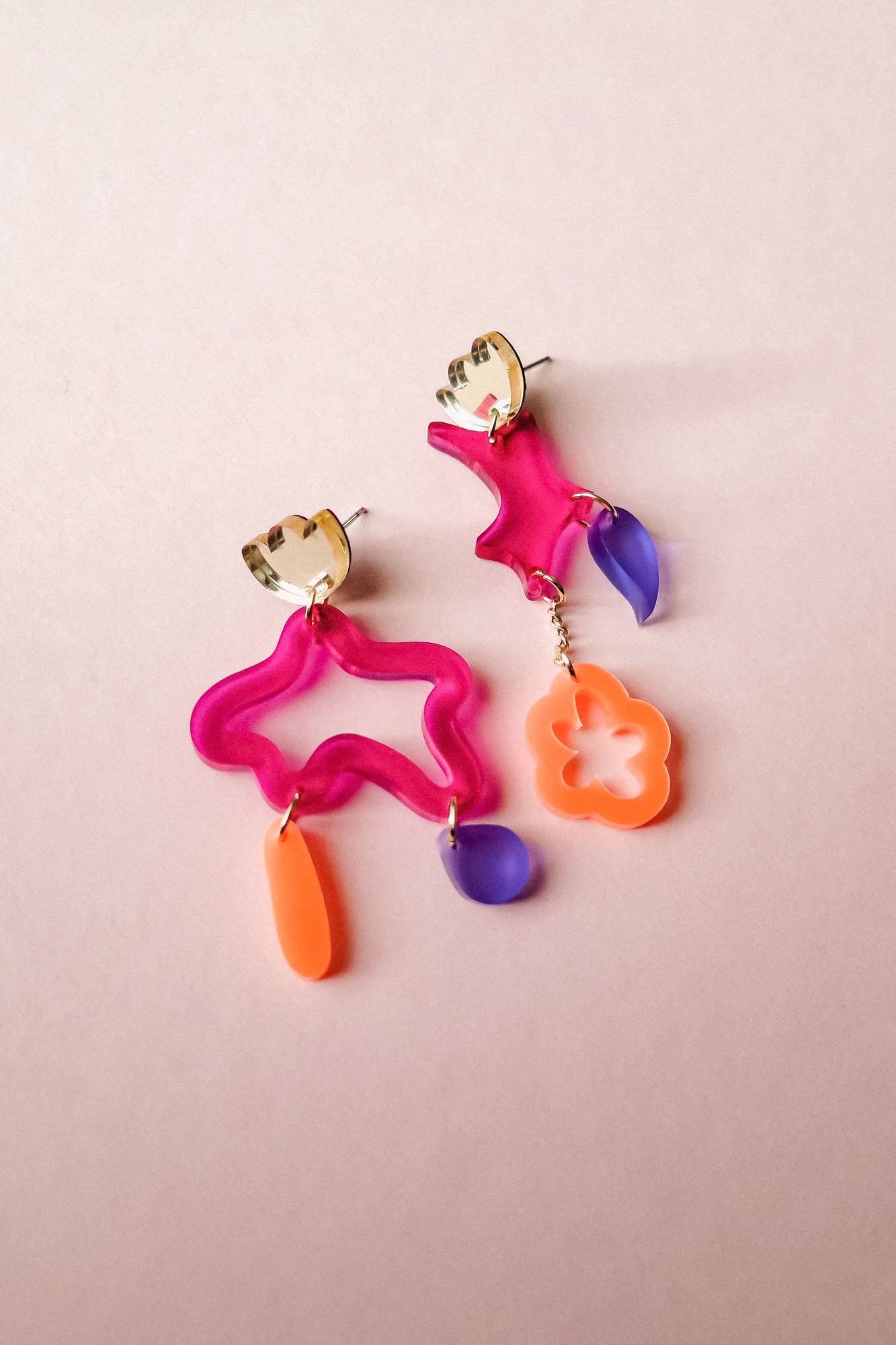 Two mismatched earrings in bold colors of pink, orange, and purple acrylic, featuring playful abstract shapes. Each unique earring dangles with geometric forms, making these Mismatched Earrings a vibrant statement on a light pink background.