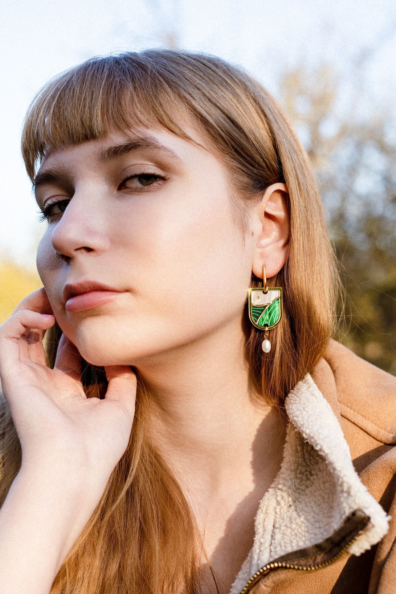 A young woman with light brown hair and bangs gazes calmly at the camera. She wears a tan fleece jacket and green and gold Art Nouveau earrings with a pearl drop. Her hand rests gently on her face, trees blurred in the background.